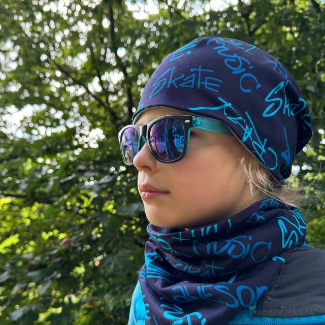 Frau trägt Wollhuhn LONG Loop, Jersey- oder Fleecefutter, Cool Music Blau, als Schal, kombiniert mit Sonnenbrille. Ideal für stilvollen Wind- und Kälteschutz.