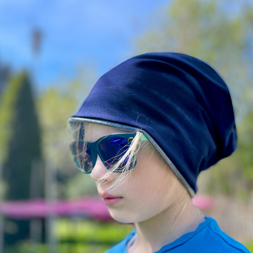 Mädchen trägt Wollhuhn Öko Long Beanie in Dunkelblau, kombiniert mit Sonnenbrille und blauem Hut, zeigt bequemen Sitz und zweilagiges Design.
