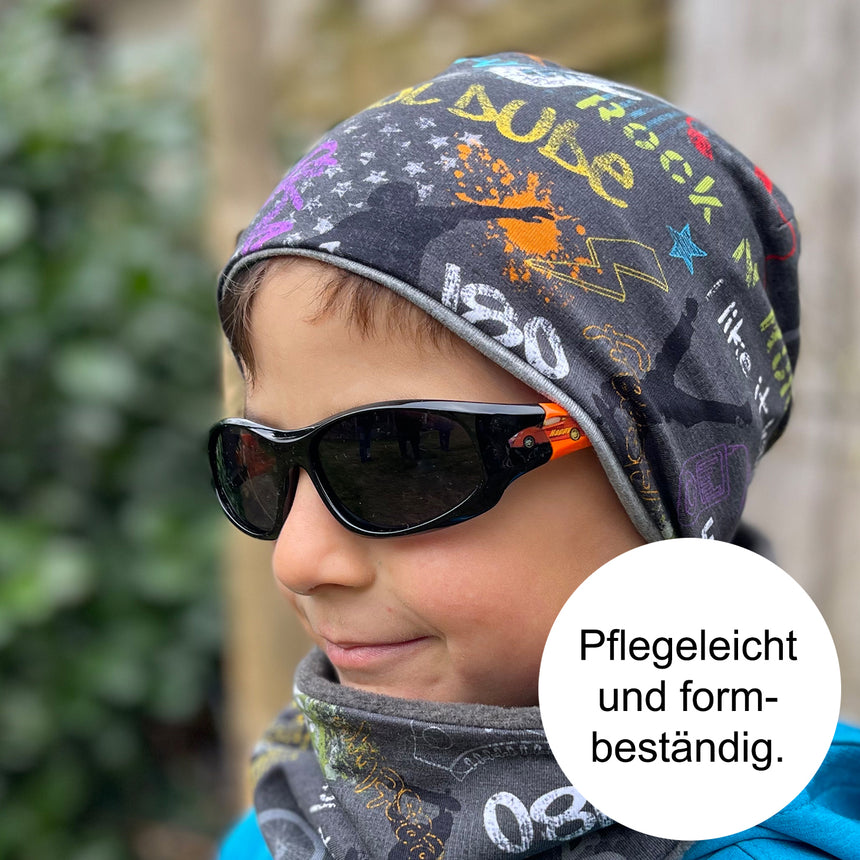 Ein Junge trägt die Wollhuhn Öko Shorties Wendemütze, zweilagig und komfortabel, kombiniert mit Sonnenbrille und Schal. Ideal für stilvolle, ganzjährige Nutzung.