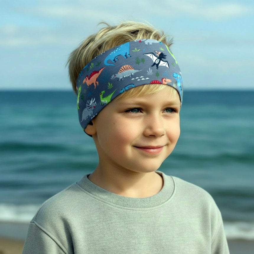 Junger Junge am Strand trägt Wollhuhn Elastisches Öko Stirnband Jersey Zweilagig Breit Dinos Grau/Bunt, lächelt leicht, Meer im Hintergrund. Ideal für Outdoor-Aktivitäten.