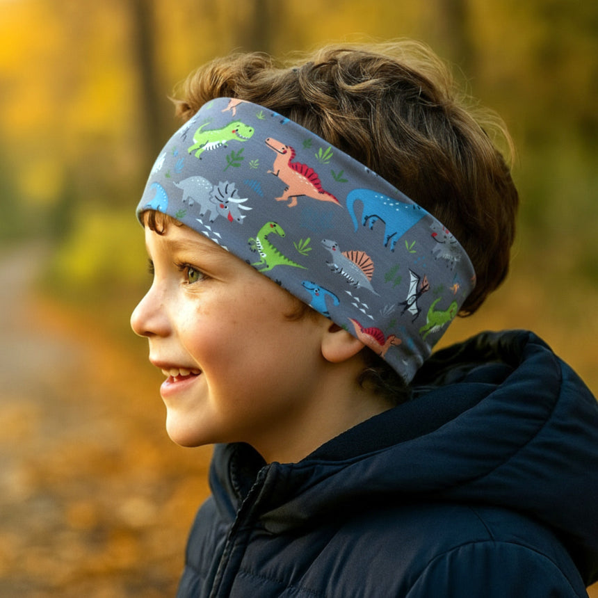 Junge mit Wollhuhn Elastisches Öko Stirnband, Jersey, zweilagig, breit, Dinos grau/bunt, lächelnd auf herbstlichem Weg, betont Weichheit und Vielseitigkeit des Stirnbands.