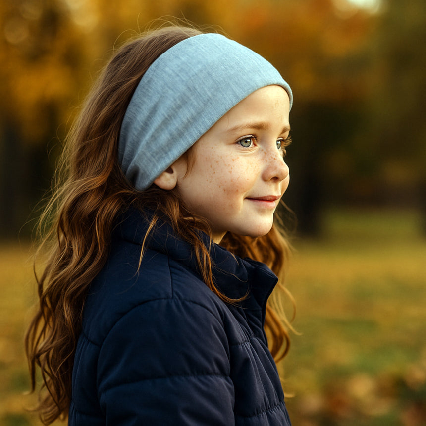 Mädchen im Profil mit Wollhuhn Elastisches Öko Stirnband in Hellgrau, trägt blaues Steppjacke, umgeben von herbstlicher Landschaft.