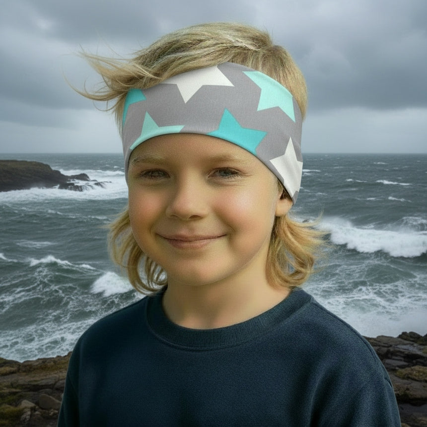 Junges Kind am felsigen Ufer trägt ein Wollhuhn Elastisches Öko Stirnband/Haarband Jersey Zweilagig Breit Big Stars Sterne Grau/Mint, ideal für Outdoor-Aktivitäten.