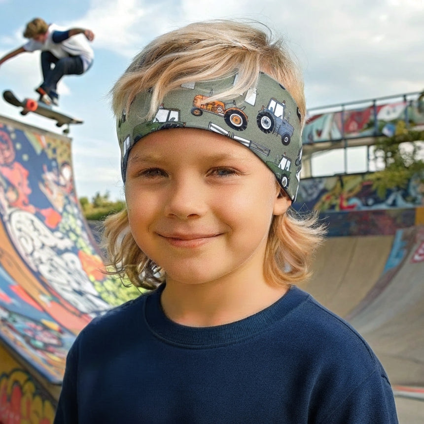 Kind mit blondem Haar trägt ein Wollhuhn Elastisches Öko Stirnband / Haarband Jersey Zweilagig Breit Traktor/Trecker Grün in einem Skatepark.