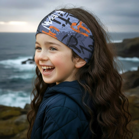 Mädchen am felsigen Ufer trägt Wollhuhn Elastisches Öko Stirnband, lächelt glücklich, während Wellen brechen. Stirnband aus weichem, zertifiziertem Bio-Jersey, vielseitig tragbar.