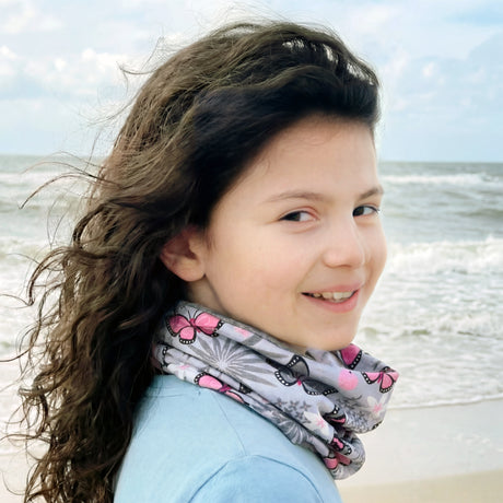 Junges Mädchen am Strand mit Wollhuhn Leichtes Lockeres Öko Jersey Halstuch, grau mit rosa Schmetterlingen, das Haar vom Wind zerzaust, lächelnd zur Kamera.