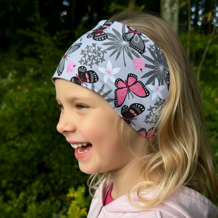 Junges Mädchen mit einem breiten Wollhuhn Elastisches Öko Stirnband, zweilagig aus Jersey mit Schmetterlingen, lächelt im Grünen. Ideal für vielseitige Outdoor-Aktivitäten.