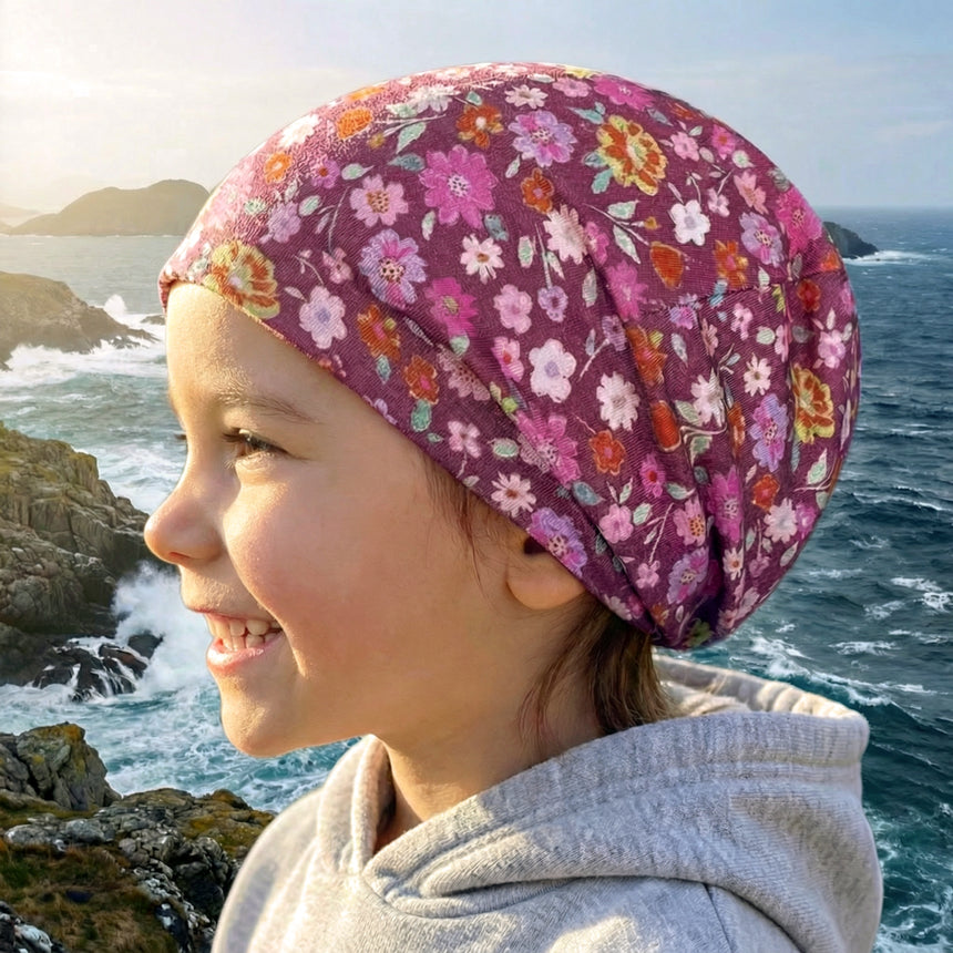 Ein Kind trägt eine Wollhuhn Öko Shortie Wendemütze in Blumen Beere, blickt lächelnd auf eine felsige Küste mit Wellen im Hintergrund.