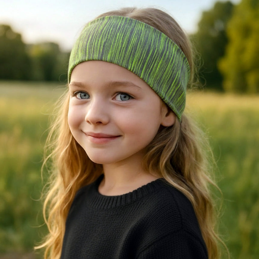 Mädchen mit blondem Haar trägt Wollhuhn Elastisches Öko Stirnband in Grüntönen, stehend auf einer Wiese; handgemacht aus nachhaltigem Bio-Jersey, ideal für Outdoor-Aktivitäten.