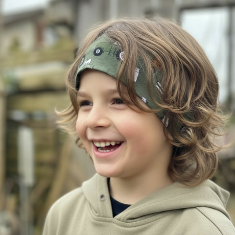 Ein Kind im Freien trägt ein Wollhuhn Elastisches Öko Stirnband, Jersey zweilagig breit, mit grünem Traktor-Muster, das Haare zurückhält.