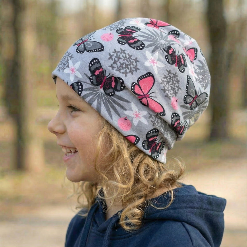 Kind seitlich im Wald, trägt Wollhuhn Öko Shorties Beanie, grau mit Schmetterlingsmuster, lächelt in dunkelblauem Hoodie; handgemacht, zweilagig, flexibel und bequem.