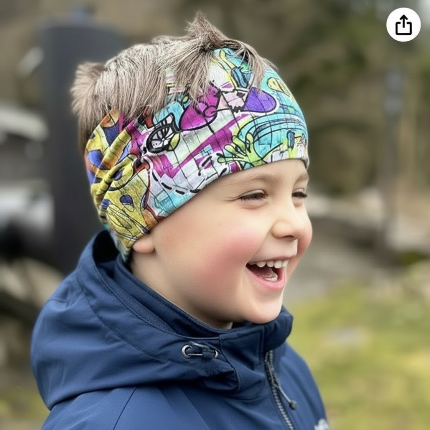 Kind mit Wollhuhn Elastisches Öko Stirnband / Haarband Jersey Zweilagig Breit Graffiti Bunt, lächelnd im Freien, trägt dunkle Jacke, Stirnband hochgeschoben.