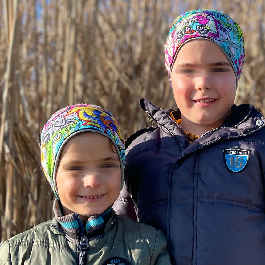 Wollhuhn Öko Long Beanie: Zwei Kinder mit bunten Beanies vor hohem Gras. Bequeme, zweilagige, wendbare Mütze, ideal für das ganze Jahr.