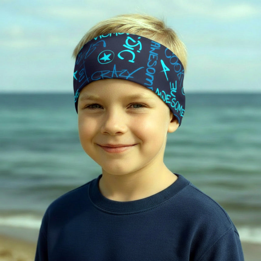 Junger Junge trägt das Wollhuhn Elastisches Öko Stirnband/Haarband Jersey Zweilagig Breit Cool Music Blau vor ruhigem Meer, lächelt leicht in die Kamera.