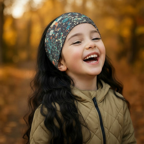 Mädchen im Herbstwald trägt ein Wollhuhn Elastisches Öko Stirnband, lacht fröhlich. Stirnband zweilagig, breit, ideal für Outdoor-Aktivitäten. Handgemacht in Deutschland.