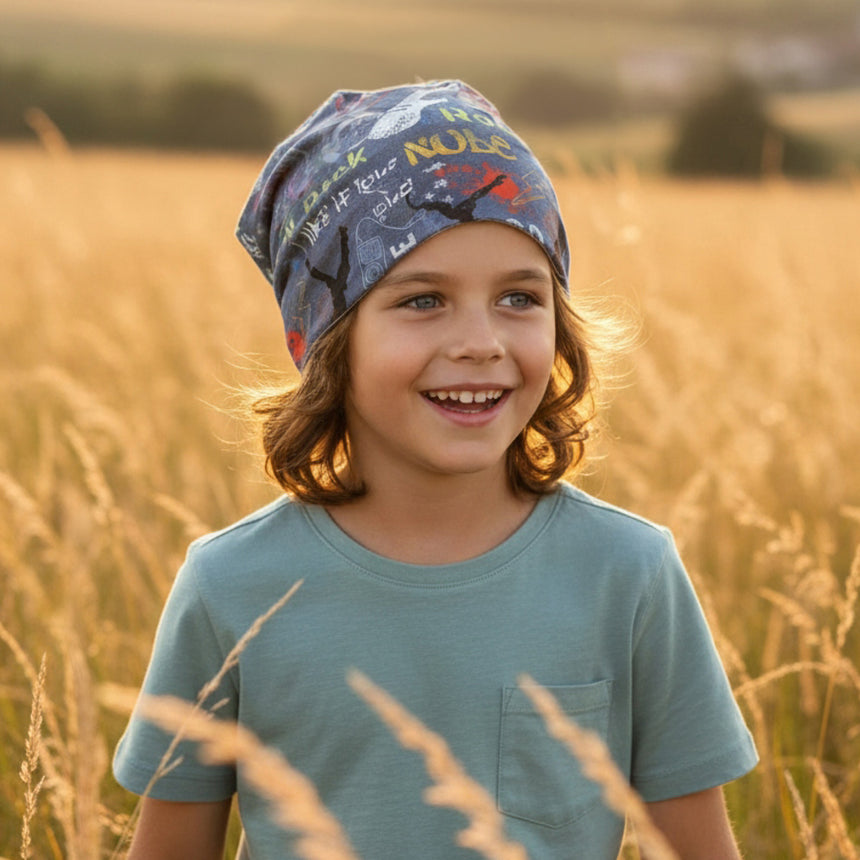 Kind mit breitem Lächeln trägt eine Wollhuhn Öko Long Beanie / Wendemütze in Jeansblau, stehend in einem sonnigen Feld mit goldenem Gras.