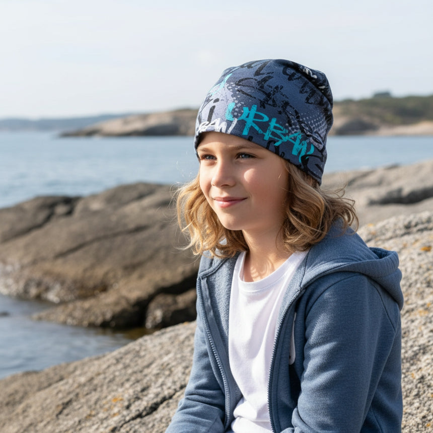Ein Kind mit blondem Haar trägt eine Wollhuhn Öko Shorties Wendemütze in Skater Blau, sitzt am Ufer und blickt aufs Wasser.