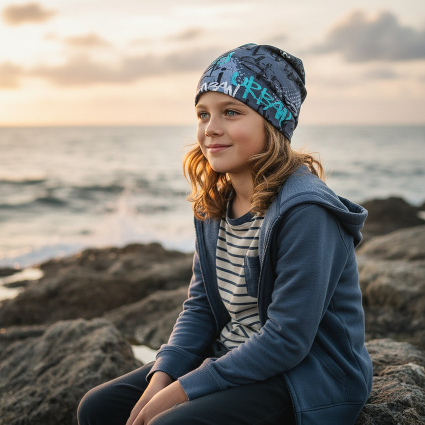 Kind mit Wollhuhn Öko Shorties Wendemütze, sitzt am felsigen Ufer bei Sonnenuntergang. Die Mütze ist handgefertigt, bequem und mitwachsend, perfekt für aktive Kinder.