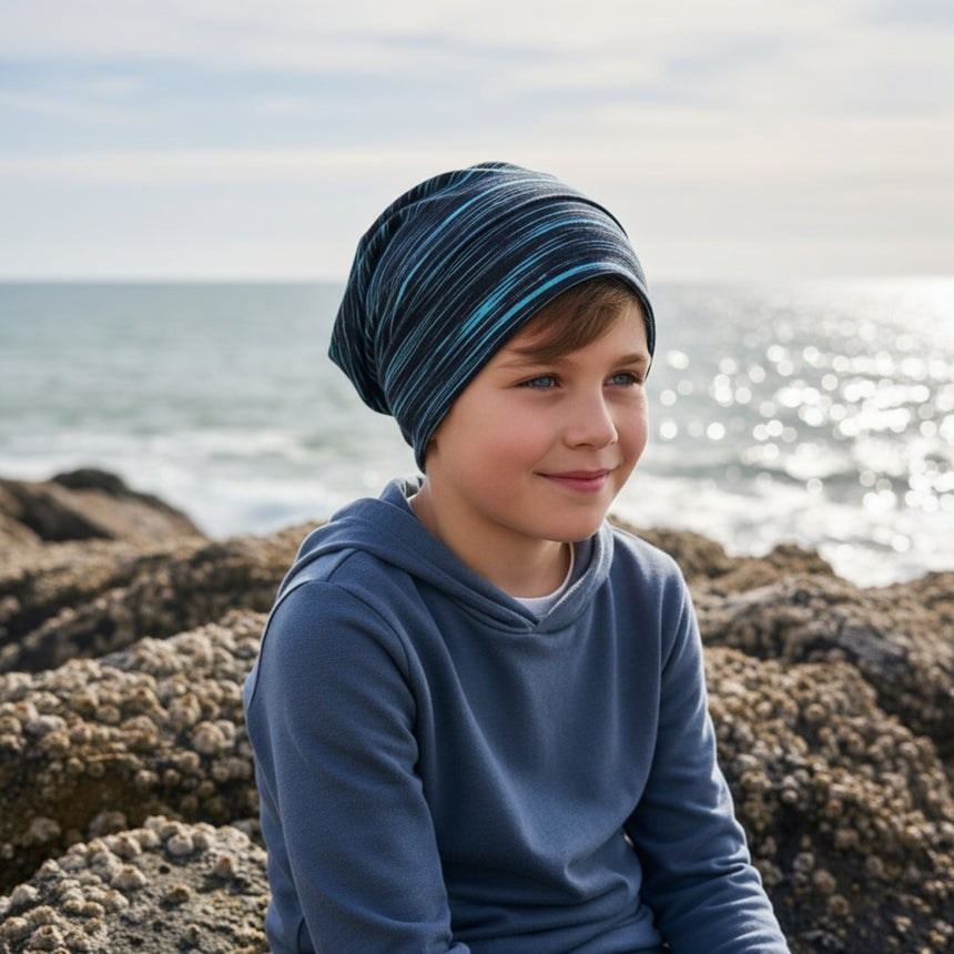 Junger Junge sitzt am Meer, trägt Wollhuhn Öko Long Beanie/Wendemütze, für Ganzjährig, zweilagig, Lines Schwarz/Türkis. Handgemacht, lässig, bequem. Ideal für Outdoor-Abenteuer.