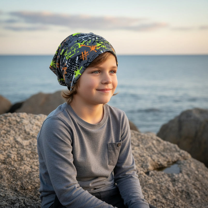 Junge sitzt am felsigen Ufer, trägt Wollhuhn Öko Long Beanie/Wendemütze, graffiti-grau/bunt. Handgemacht, lässiger Look, ideal für Frühling und milde Wintertage.