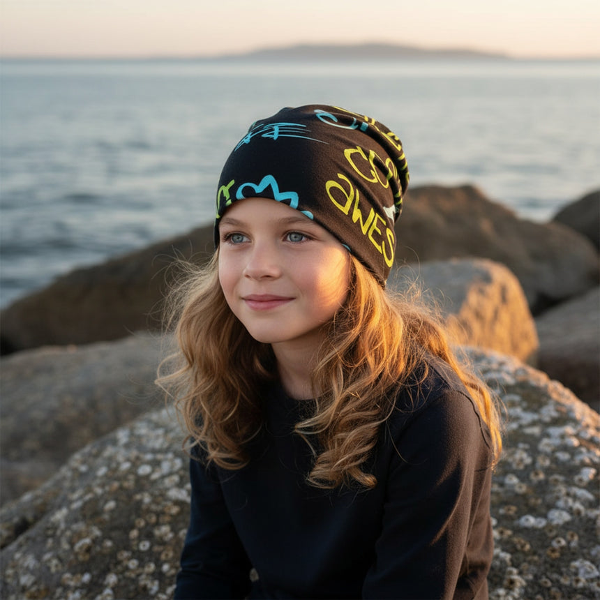 Ein Mädchen trägt die Wollhuhn Öko Shorties / Wendemütze am Strand, zeigt die zweilagige, dehnbare Beanie in Schwarz mit bunten Mustern bei Sonnenuntergang.