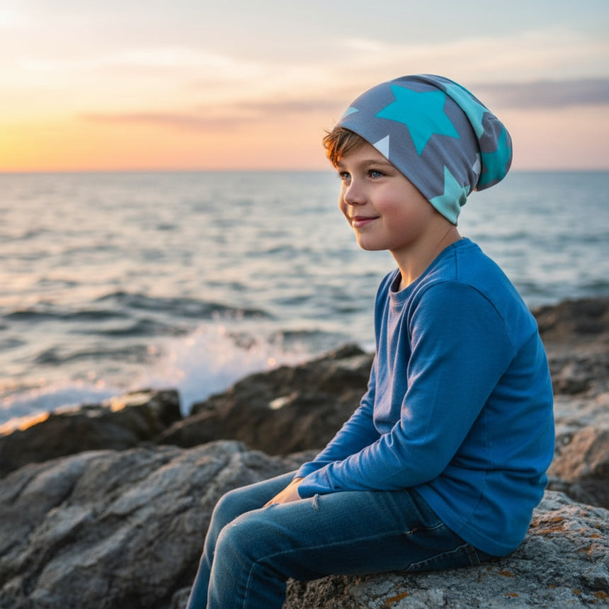 Junge am Felsen trägt Wollhuhn Öko Long Beanie mit großen Sternen, blickt auf Sonnenuntergang am Meer. Zweilagig, bequem, ideal für das ganze Jahr.