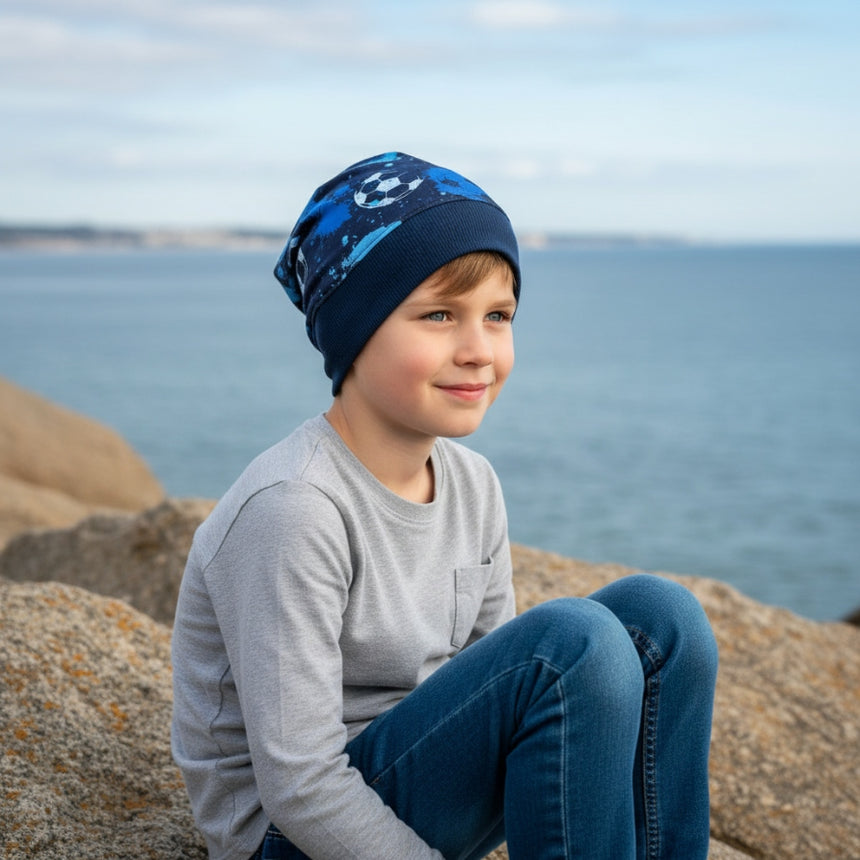 Junge trägt Wollhuhn Dünne Öko Beanie-Mütze Sommer/Übergang Einlagig Fußball Blautöne, sitzt auf Felsen am Wasser, betont handgemachte, nachhaltige Qualität.