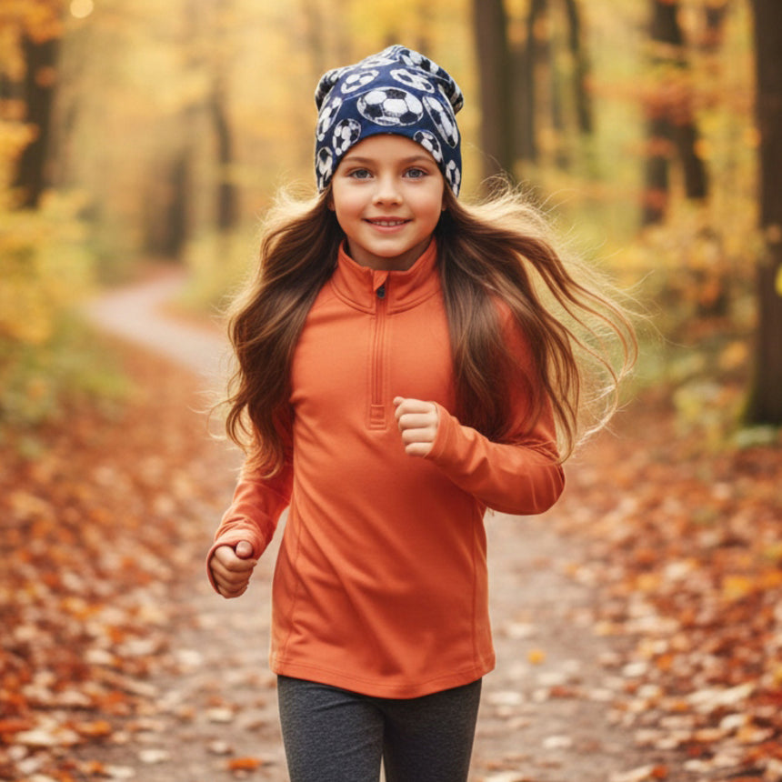 Mädchen joggt auf Herbstwaldweg, trägt Wollhuhn Öko Long Beanie/Wendemütze, zweilagig, fußballgemustert, für lässigen Look und Komfort, ideal für milde Tage.