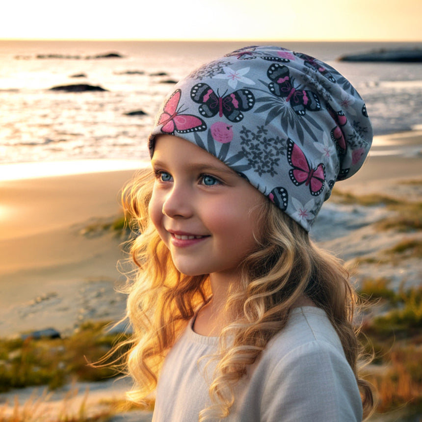 Mädchen am Strand bei Sonnenuntergang trägt Wollhuhn Öko Long Beanie, Wendemütze, zweilagig mit Schmetterlingen, lässig am Hinterkopf fallend, ideal für Kinder.