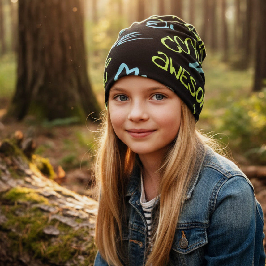 Mädchen trägt Wollhuhn Öko Shorties Wendemütze mit buntem Graffiti-Design, sitzt in sonnigem Wald. Bequeme, zweilagige Beanie, ideal für das ganze Jahr.