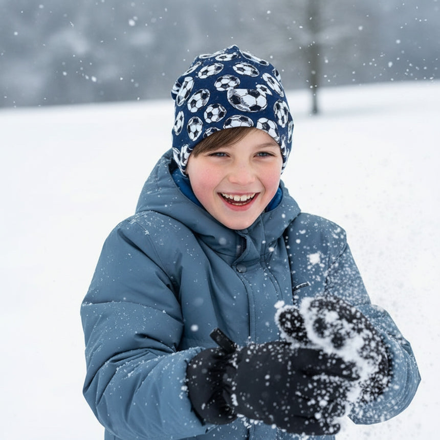 Kind im Schnee mit Wollhuhn Öko Shortie, Wendemütze, Fußball Dunkelblau, lächelnd und Schnee werfend, zeigt die bequeme und stylische, zweilagige Mütze für aktive Kinder.