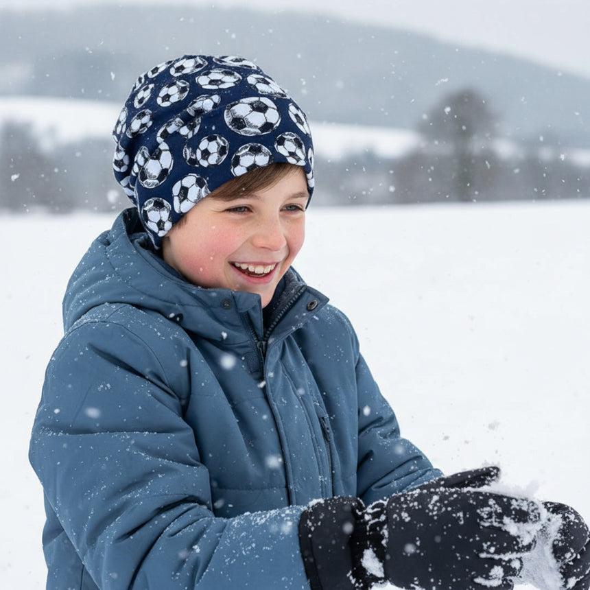 Junge im Schnee mit Wollhuhn Öko Long Beanie, Wendemütze, Fußball Dunkelblau. Lächelnd, trägt blaue Jacke und formbare Schneehandschuhe. Perfekt für milde Wintertage.