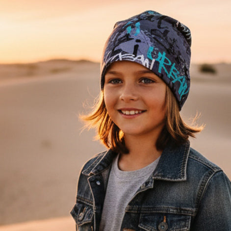 Kind im grauen Wollhuhn Öko Shorties Beanie, lächelt sanft in der Wüste bei Sonnenuntergang. Die Wendemütze bietet lässigen Stil und Komfort für aktive Kinder.