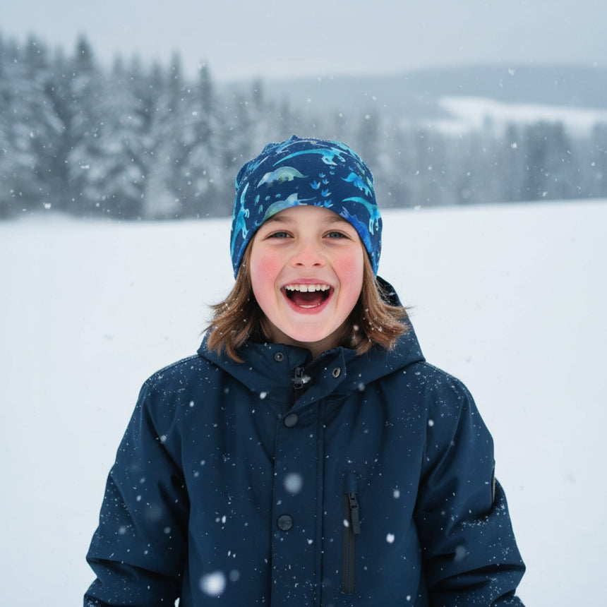Kind im Schnee mit Wollhuhn Öko Long Beanie, Dinos Blau, lächelt. Zweilagige Wendemütze bietet Komfort und Wärme, ideal für Freizeit und mildere Wintertage.