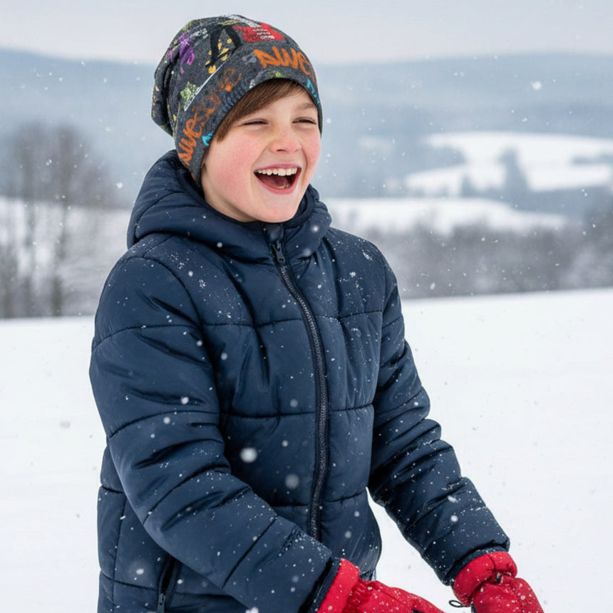 Ein Junge im Schnee trägt die Wollhuhn Öko Long Beanie, handgemacht, zweilagig, wendbar, perfekt für milde Wintertage. Er lacht, umgeben von fallenden Schneeflocken.