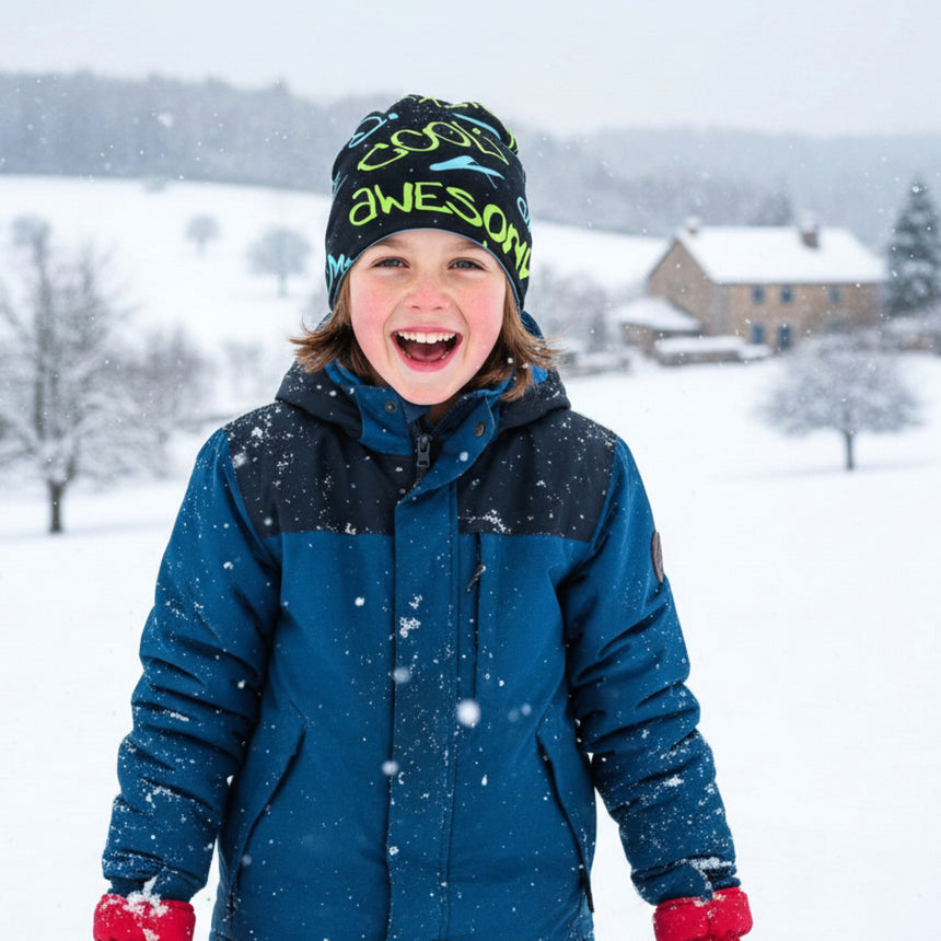 Kind im Schnee mit Wollhuhn Öko Long Beanie, lässig sitzend. Handgemachte, zweilagige Wendemütze für Kinder, ideal für mildes Winterwetter. Grinsend, vor Bauernhaus und Bäumen.