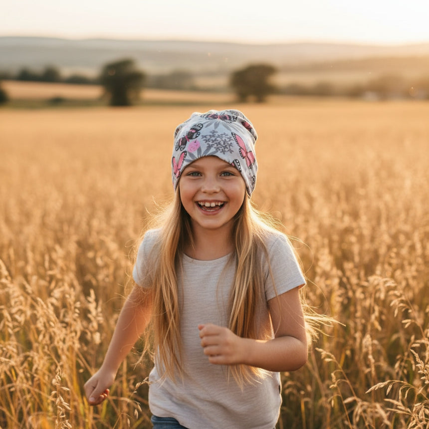 Mädchen mit Wollhuhn Öko Long Beanie, Schmetterlinge Grau/Rosa, läuft lächelnd durch goldenes Feld. Handgemachte, zweilagige Wendemütze, ideal für alle Jahreszeiten.
