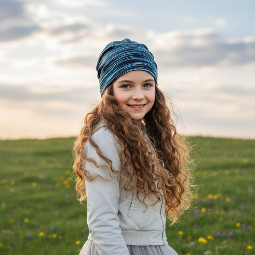 Mädchen mit Wollhuhn Öko Shorties Beanie, zweilagig, in Schwarz/Türkis gestreift, lächelt in blumiger Wiese; handgemacht, bequem und ideal für aktive Kinder.