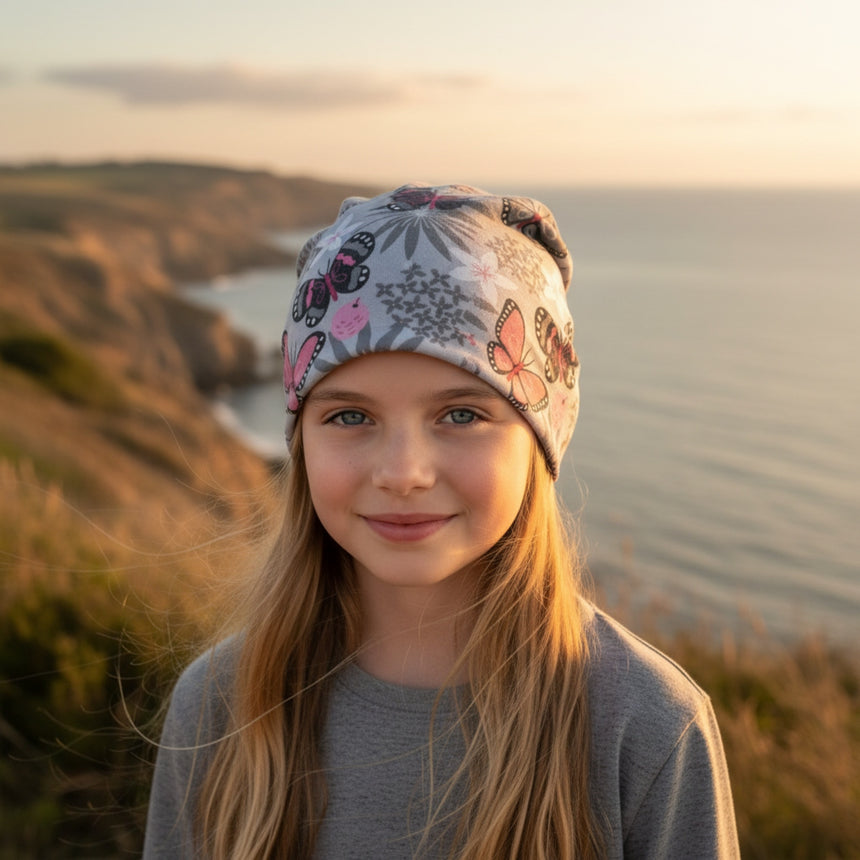 Ein Mädchen trägt eine Wollhuhn Öko Shorties Wendemütze mit Schmetterlingen, steht auf einer Klippe mit Meerblick, lächelt sanft zur Kamera.