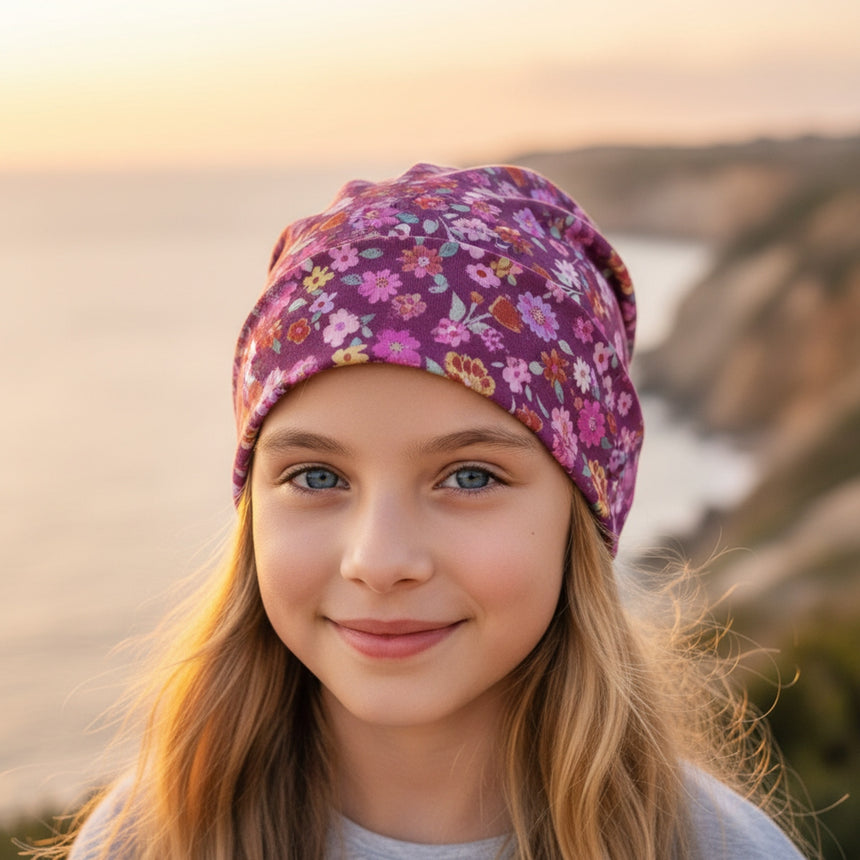 Mädchen mit Blumen-Beanie von Wollhuhn, lächelt vor Küstenlandschaft. Handgefertigte, zweilagige Öko-Wendemütze, ideal für Kinder.