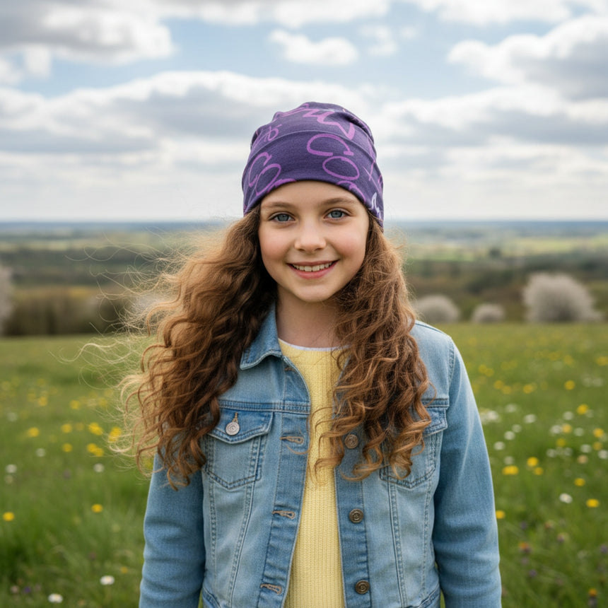 Mädchen trägt Wollhuhn Öko Long Beanie in violett/beere, lässig in einer blühenden Wiese stehend, passend für Frühling und Herbst, handgemacht und zweilagig.