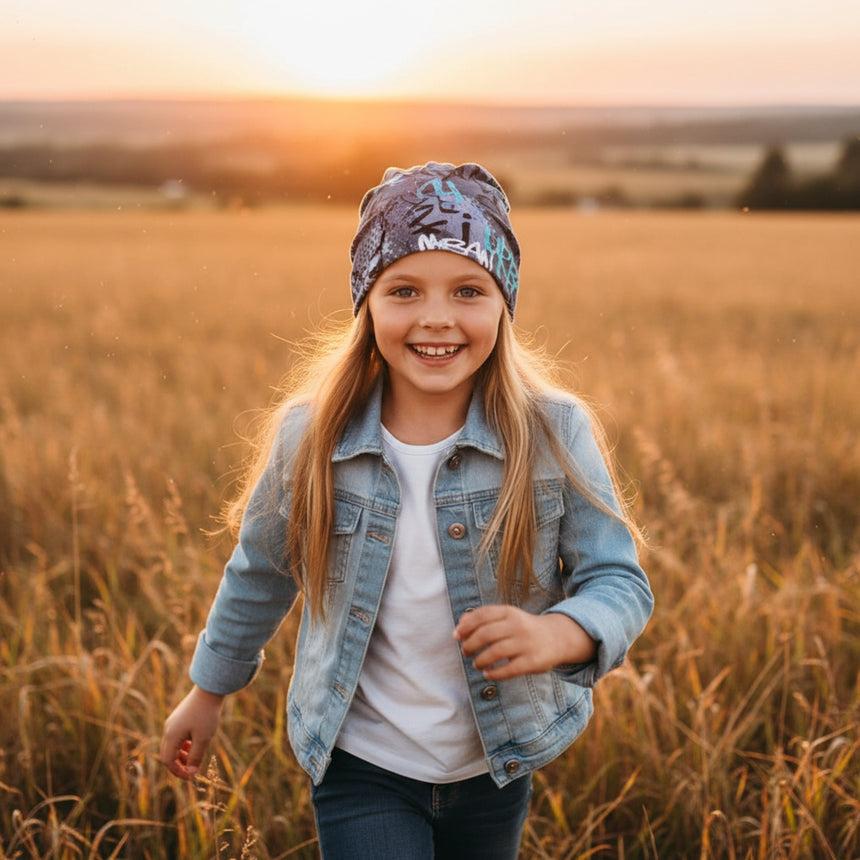Mädchen mit Wollhuhn Öko Shorties Beanie, lässig und bequem, rennt lächelnd durch ein goldener Feld bei Sonnenuntergang, ideal für aktive Kinder.