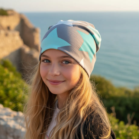 Mädchen trägt Wollhuhn Öko Shorties Wendemütze, zweilagig, große Sterne grau/mint, am Meer. Handgemacht, weich, für Kinder.