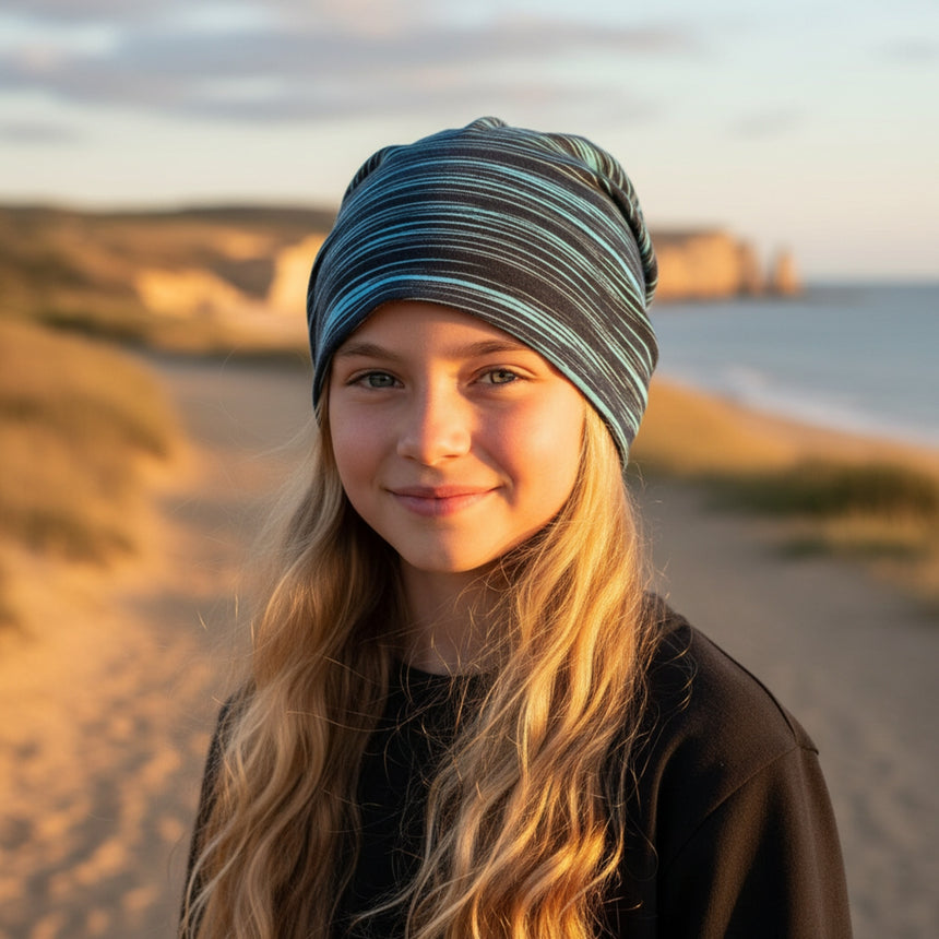 Mädchen am Strand trägt Wollhuhn Öko Shorties, eine zweilagige, ganzjährig nutzbare Wendemütze in Lines Schwarz/Türkis, passend für aktive Kinder.