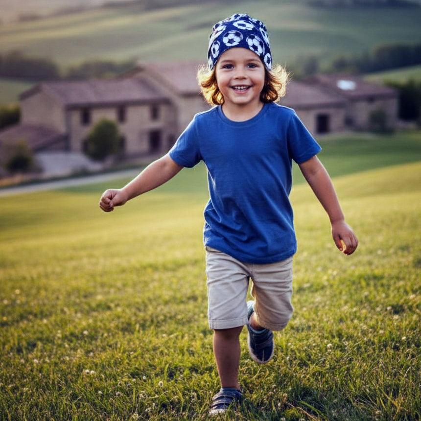 Junger Junge trägt Wollhuhn Öko Shorties Wendemütze Fußball Dunkelblau, läuft fröhlich über eine Wiese. Ideal für aktive Kinder, handgemacht und mitwachsend.