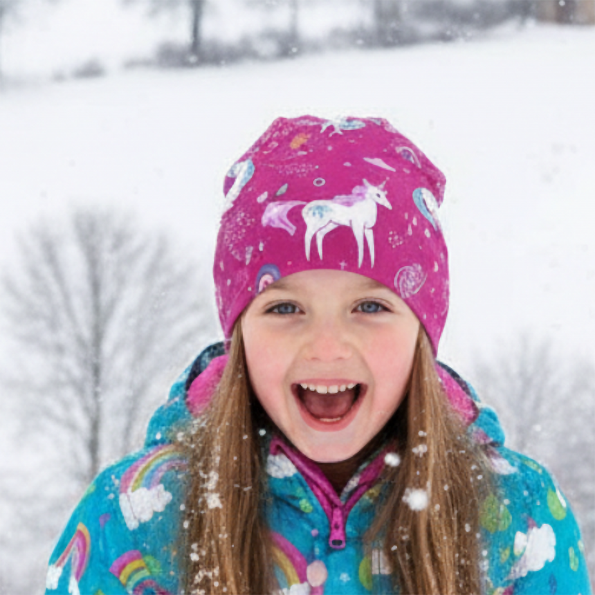 Mädchen im Schnee trägt Wollhuhn Öko Shorties Wendemütze mit Einhörner-Beere-Design, passend und wärmend für aktive Kinder. Sie lacht und genießt die Winterlandschaft.