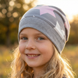 Kind mit langen, welligen blonden Haaren trägt eine Wollhuhn Dünne Öko Beanie-Mütze Sommer/Übergang Einlagig Big Stars Sterne Grau/Rosa/Pink und lächelt sanft in die Kamera.