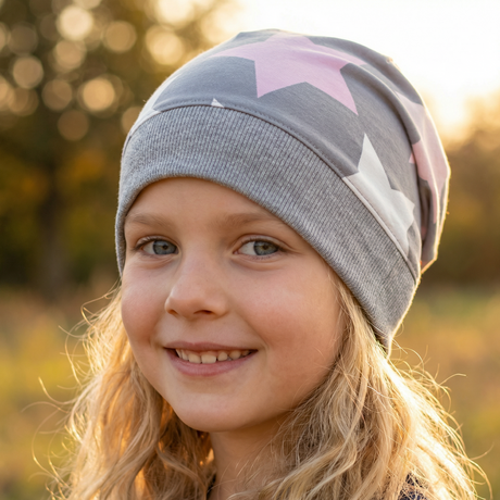 Kind mit langen, welligen blonden Haaren trägt eine Wollhuhn Dünne Öko Beanie-Mütze Sommer/Übergang Einlagig Big Stars Sterne Grau/Rosa/Pink und lächelt sanft in die Kamera.