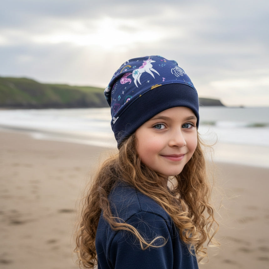 Mädchen am Strand trägt Wollhuhn Dünne Öko Beanie-Mütze Sommer/Übergang Einlagig mit Einhörnern in Dunkelblau, hergestellt aus weichem, nachhaltigem Öko-Jersey.