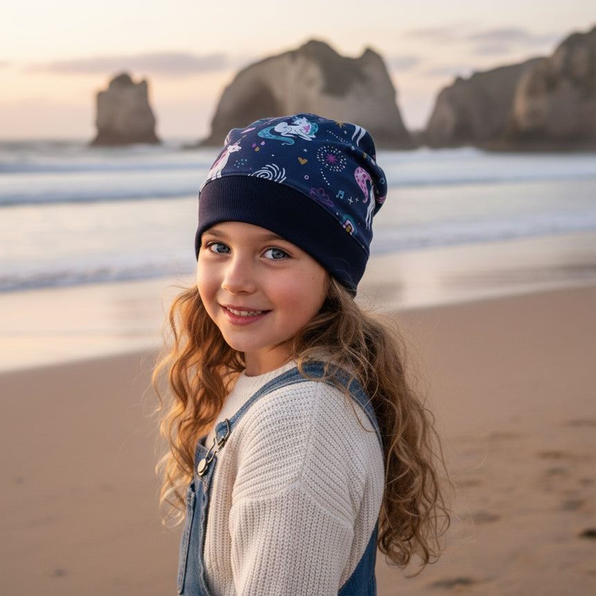 Mädchen am Strand trägt Wollhuhn Dünne Öko Beanie-Mütze Sommer/Übergang Einlagig Einhorn, lächelt in die Kamera. Handgemacht, dehnbar, ideal für kühle Tage.